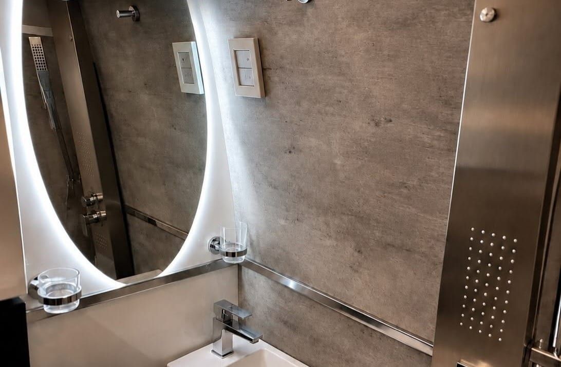 Baño con espejo ovalado iluminado, revestimiento símil cemento y columna de ducha de acero