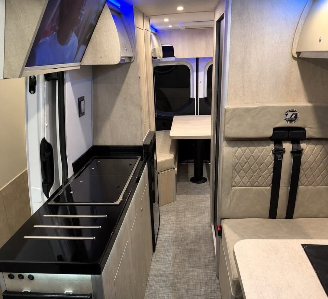 Pasillo de la Sprinter: cocina con mesada negra y TV a la izquierda, butaca doble a la derecha y el comedor al fondo